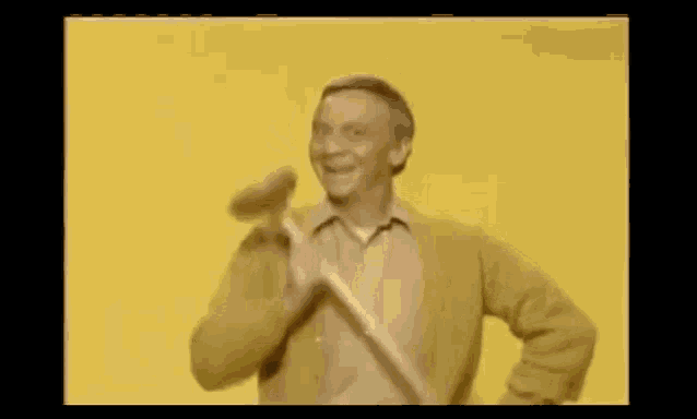 a man in a yellow sweater is holding a hammer and the name norman fell is on the screen .