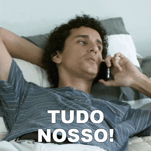 a man laying on a bed talking on a cell phone with tudo nosso written on his shirt