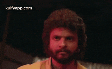 a man with a beard and curly hair is wearing a yellow shirt and looking at the camera .