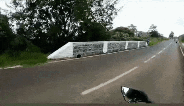 a motorcycle is driving down a road with a stone wall on the side of the road