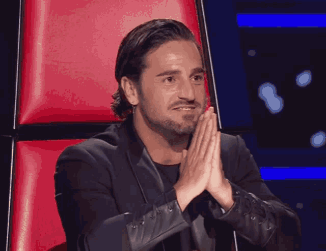 a man in a black suit sits in a red chair with his hands together