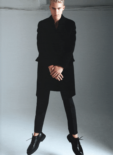 a man in a black coat and black pants is standing in the air with his hands behind his back