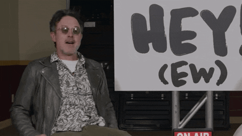 a man in a leather jacket sits in front of a sign that says hey ( ew )