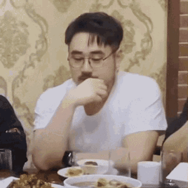 a man wearing glasses and a white shirt is sitting at a table with plates of food .