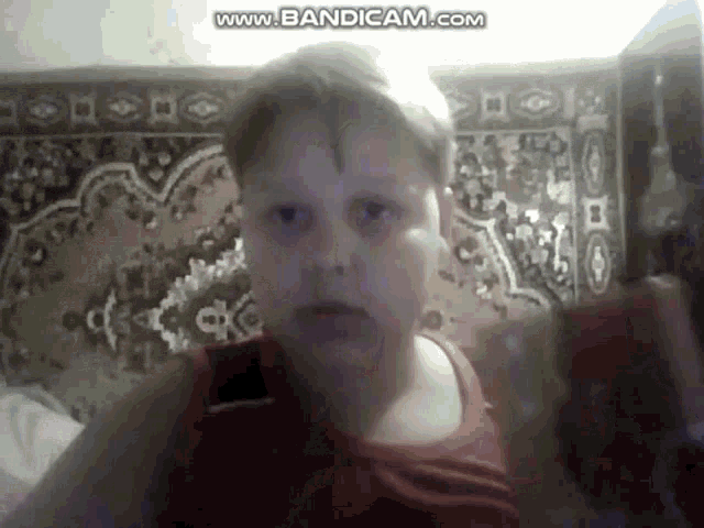 a young boy in a red tank top is sitting in front of a rug and looking at the camera .