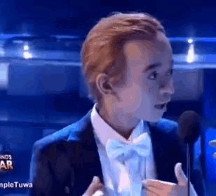 a young man in a tuxedo and bow tie is speaking into a microphone