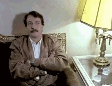 a man with a mustache sits with his arms crossed next to a lamp