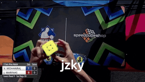 a person is holding a yellow cube in front of a speedcubeshop sign
