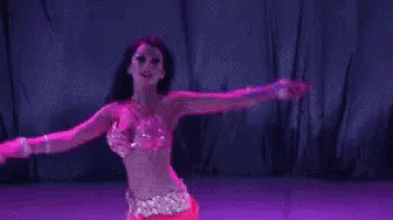 a belly dancer in a pink outfit is dancing on a stage with purple lights behind her .