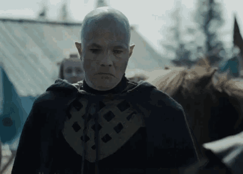 a bald man with a tattoo on his forehead stands in front of a horse and tent