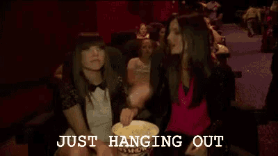 two women are sitting in a movie theater eating popcorn with the words just hanging out above them