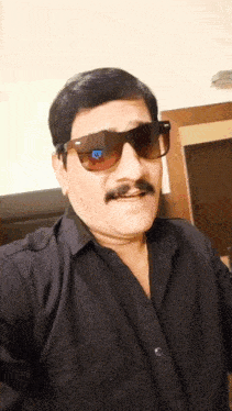 a man with a mustache wearing sunglasses and a black shirt looks at the camera