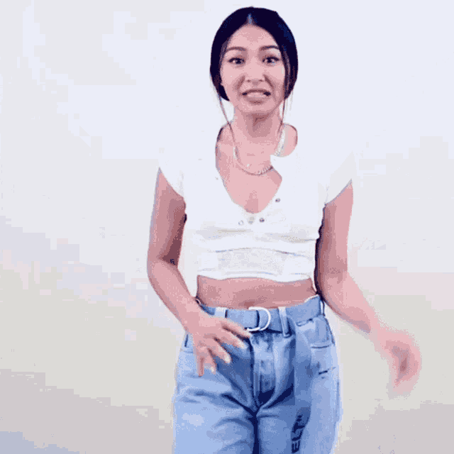 a woman wearing a white crop top and blue jeans is standing in front of a white wall .