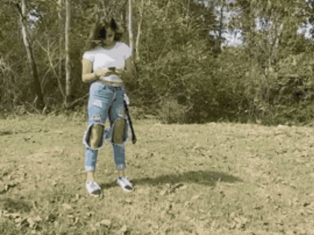 a woman wearing ripped jeans and a white shirt stands in a field looking at her phone