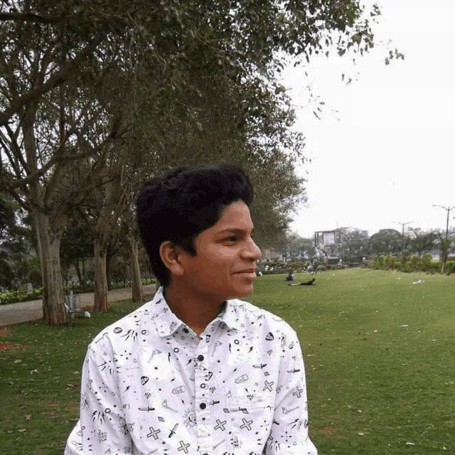 a young man wearing a white shirt with crosses on it stands in a grassy park
