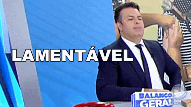 a man in a suit and tie is sitting in front of a sign that says " lamentável "