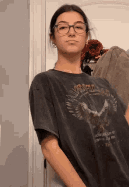 a girl wearing glasses and a t-shirt is standing in a room .