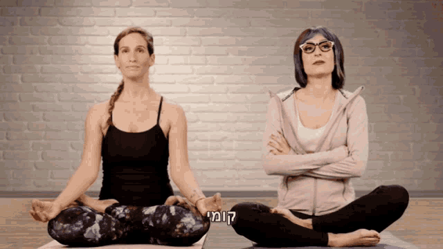 two women are sitting on yoga mats with their arms crossed in front of a brick wall and one of them has blue hair