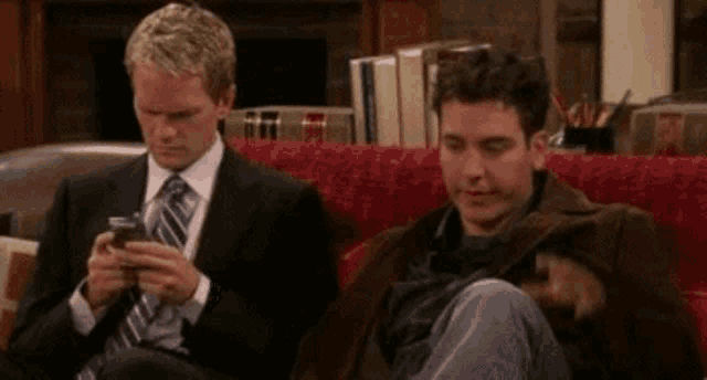 a man in a suit and tie is sitting on a couch while another man looks at his cell phone .