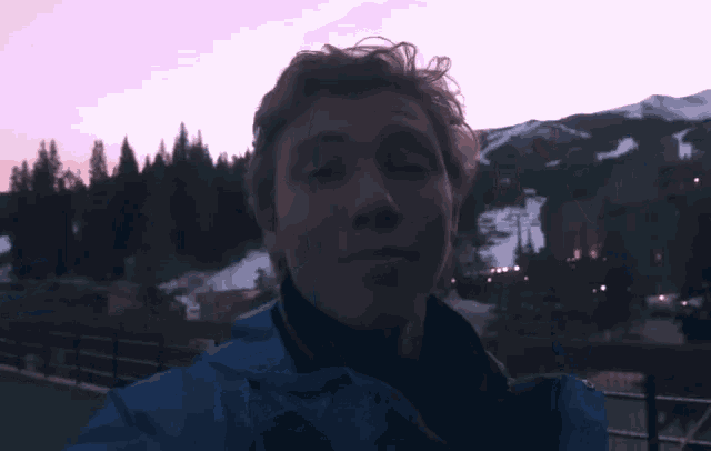 a man in a blue jacket adjusts his hair in front of a mountain