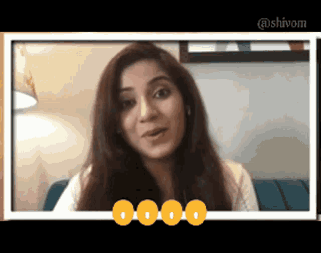 a woman is sitting on a blue couch and talking on a video call with a shivam logo in the corner