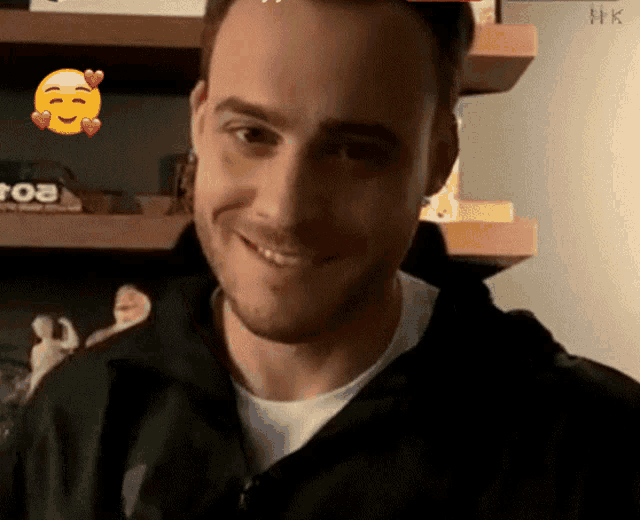 a man in a black jacket smiles in front of a shelf that says ' hk ' on it