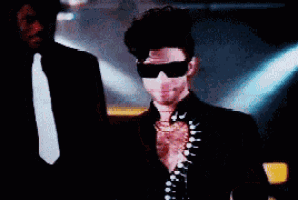 a man in a suit and tie adjusts his sunglasses while another man stands behind him