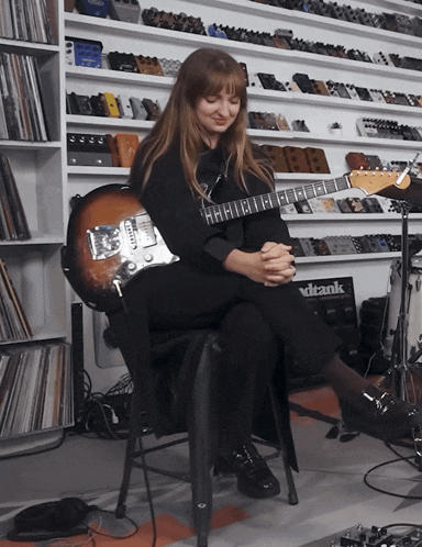 a woman sits in a chair playing a guitar in front of a deank amp