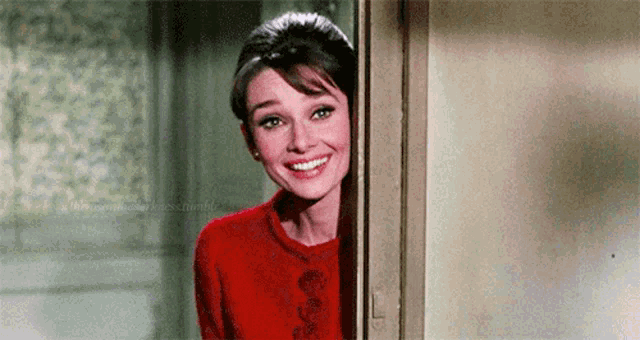 a woman in a red sweater is peeking out from behind a door and smiling .