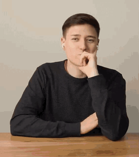 a man in a black sweater is sitting at a table with his hand on his chin .