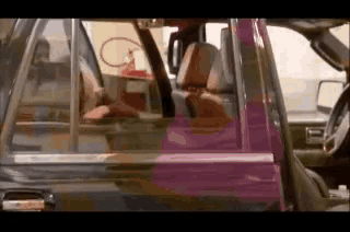 a man is driving a pink car with the door open and a woman is sitting in the back seat .