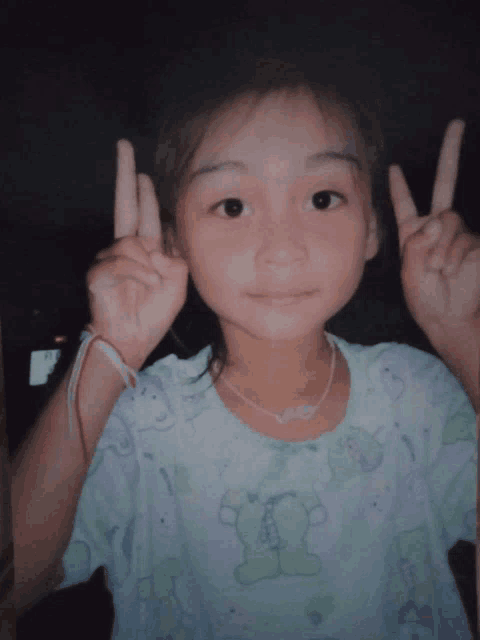 a little girl wearing a shirt with a teddy bear on it is giving a peace sign