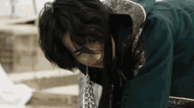 a man with long hair is wearing a green jacket and a chain around his neck .