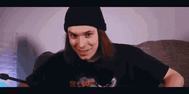 a man with long hair is sitting on a couch wearing a beanie and a black shirt .