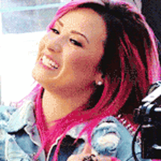 a woman with pink hair wearing a denim jacket is smiling .
