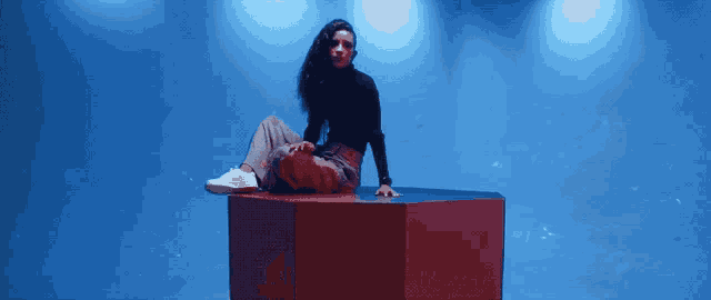 a woman sits on a red cube in front of a blue wall