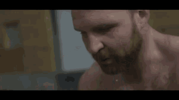 a shirtless man with a beard is standing in front of a car in a garage .