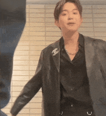 a young man wearing a black shirt and a black leather jacket is standing in front of a tiled wall .