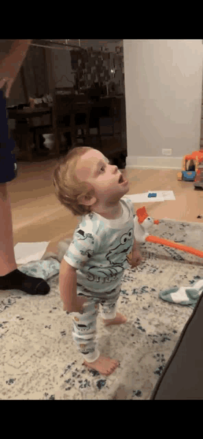 a baby in a cookie monster shirt is standing on a rug looking up .