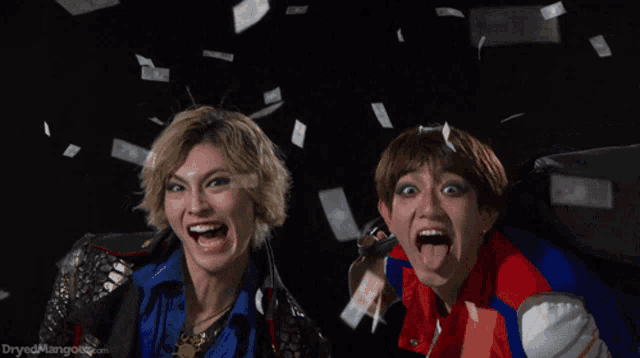 two women are standing in front of a pile of money and dryedmangoez.com is visible in the bottom right corner