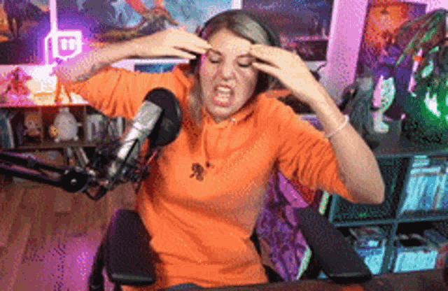 a woman in an orange hoodie is sitting in front of a microphone with her hands on her forehead .