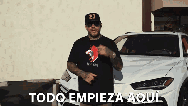 a man wearing a black shirt that says " first song " stands in front of a white car