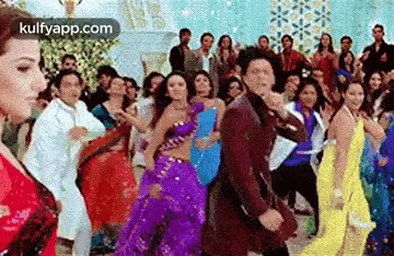 a group of people are dancing in a room with a man in a suit and a woman in a purple dress .