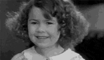 a black and white photo of a little girl with curly hair smiling and looking at the camera .