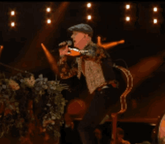 a man singing into a microphone on a stage with flowers in the background