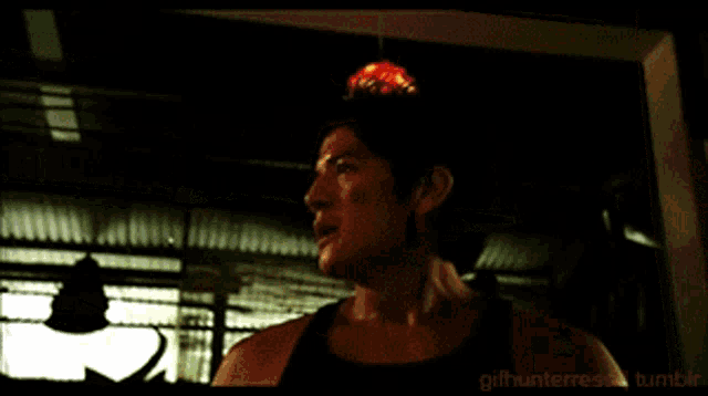 a man in a black tank top is standing in a dark room with a lamp above him