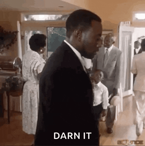 a man in a suit is standing in a room with a group of people and says darn it .