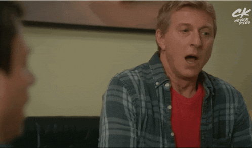 a man in a plaid shirt and a red shirt is laughing with a ck logo in the background