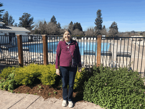 a woman in a purple jacket stands in front of a fence with a pool in the background