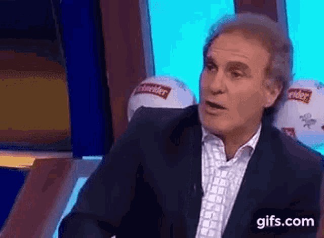 a man in a suit is talking on a television show while sitting in front of a window .